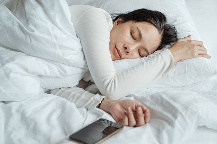 девушка брюнетка спит в кровати с белым постельным бельём, рядом лежит телефон
